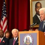 Bill Clinton speaking at a podium with audience.