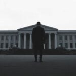A person stands solemnly, facing a distant, imposing structure.