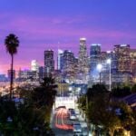 Dusk city skyline with tall illuminated buildings, palm trees in the foreground, and a residential street with red car light trails moving through the block.