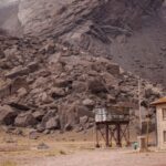 Rugged quarry landscape with a pile of rocks, a small stone building, and a metal elevated structure in a barren yard.
