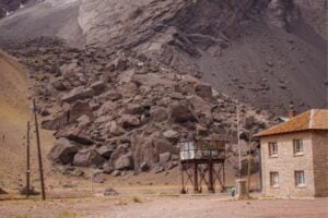 Rugged quarry landscape with a pile of rocks, a small stone building, and a metal elevated structure in a barren yard.