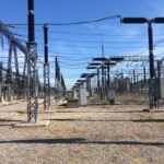 High-voltage power substation with metal lattice towers, insulators, and long rows of electrical equipment across a gravel lot.
