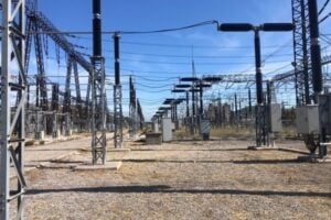 High-voltage power substation with metal lattice towers, insulators, and long rows of electrical equipment across a gravel lot.