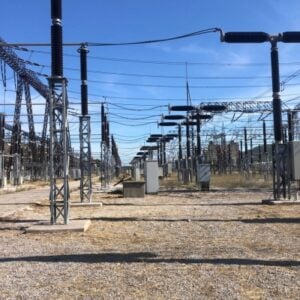High-voltage power substation with metal lattice towers, insulators, and long rows of electrical equipment across a gravel lot.