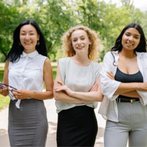 Three women stand together on a sunny park path, smiling at the camera, one holding papers.