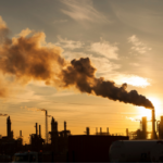 Industrial refinery at sunset with a dark smoke plume rising from a tall chimney against an orange sky.