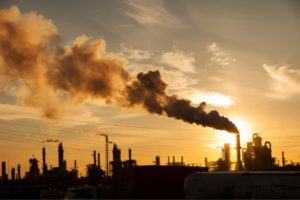 Industrial refinery at sunset with a dark smoke plume rising from a tall chimney against an orange sky.