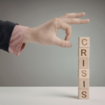 Hand hovering over a vertical stack of wooden letter blocks spelling CRISIS on a table.