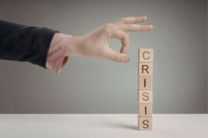 Hand hovering over a vertical stack of wooden letter blocks spelling CRISIS on a table.