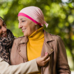 Two women chatting outdoors in a park, one adjusting the other’s leopard scarf as they smile at each other, wearing a brown leather jacket and a mustard turtleneck with a pink headscarf, outstretched greenery in the background.