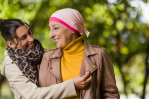 Two women chatting outdoors in a park, one adjusting the other’s leopard scarf as they smile at each other, wearing a brown leather jacket and a mustard turtleneck with a pink headscarf, outstretched greenery in the background.
