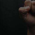 Close-up of a clenched fist against a dark background, showing knuckles and fingers curled in.