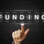 Hand with index finger pointing at digital flip panels spelling FUNDING on a dark background.