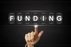 Hand with index finger pointing at digital flip panels spelling FUNDING on a dark background.