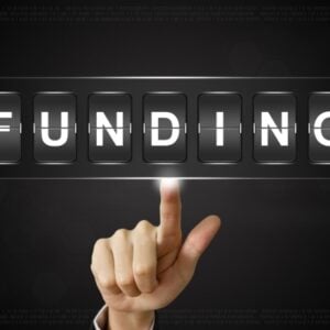 Hand with index finger pointing at digital flip panels spelling FUNDING on a dark background.