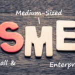 Wooden letters spell 'SME' on a wooden surface, with handwritten notes 'Medium-Sized' above and 'Small & Enterprises' beside—illustrating the SME acronym.