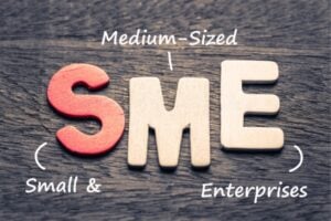 Wooden letters spell 'SME' on a wooden surface, with handwritten notes 'Medium-Sized' above and 'Small & Enterprises' beside—illustrating the SME acronym.