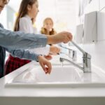 Children washing hands at a white school sink, turning taps with faucets on, in a classroom bathroom setting in the background
