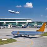 Airplane with a yellow tail taxiing on a runway near a modern terminal; another jet lifts off in the blue sky behind it.