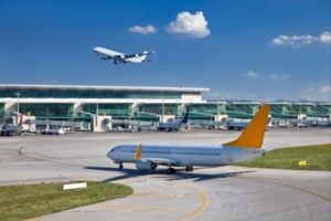 Airplane with a yellow tail taxiing on a runway near a modern terminal; another jet lifts off in the blue sky behind it.