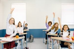 Classroom scene with smiling students raising hands at their desks, backpacks and apples visible on desks in a bright room.