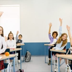 Classroom scene with smiling students raising hands at their desks, backpacks and apples visible on desks in a bright room.