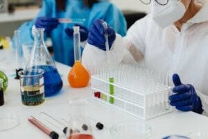 Scientist wearing a mask and gloves uses a pipette to transfer liquid into a test tube rack in a bright lab.