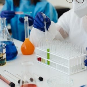 Scientist wearing a mask and gloves uses a pipette to transfer liquid into a test tube rack in a bright lab.