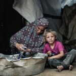 A woman in a patterned headscarf washes clothes in a large tub while a young girl sits beside her in a shelter made of blankets.