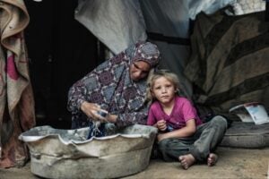 A woman in a patterned headscarf washes clothes in a large tub while a young girl sits beside her in a shelter made of blankets.
