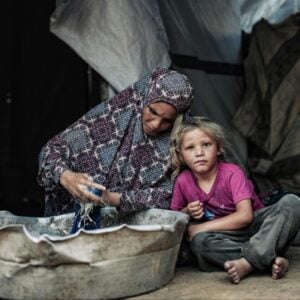 A woman in a patterned headscarf washes clothes in a large tub while a young girl sits beside her in a shelter made of blankets.
