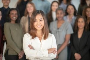 Front-center woman in a white blouse with arms crossed, smiling, standing in front of a diverse group of colleagues.