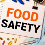 Notebook page reading 'FOOD SAFETY' with gloves, a mask, thermometer and pills on a desk-filled safety setup.