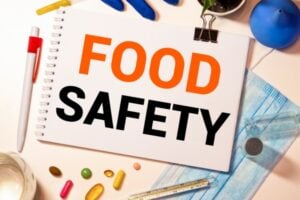 Notebook page reading 'FOOD SAFETY' with gloves, a mask, thermometer and pills on a desk-filled safety setup.