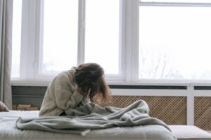 A person sits on a bed in a bright bedroom, head in hands, wrapped in a blanket, looking distressed or sad.