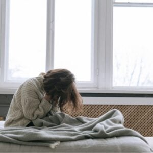 A person sits on a bed in a bright bedroom, head in hands, wrapped in a blanket, looking distressed or sad.