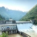 Hydropower dam in a green valley with spillways pouring water and a small control building, mountains in the background.