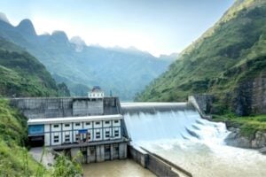 Hydropower dam in a green valley with spillways pouring water and a small control building, mountains in the background.