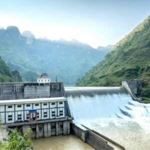 Hydropower dam in a green valley with spillways pouring water and a small control building, mountains in the background.