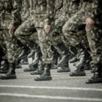 Soldiers in camouflage uniforms marching in formation, showing their boots and lower bodies in step.
