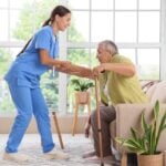 Caregiver in blue scrubs assists an elderly man with a walking cane, helping him stand in a bright living room.