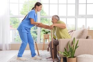 Caregiver in blue scrubs assists an elderly man with a walking cane, helping him stand in a bright living room.