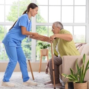 Caregiver in blue scrubs assists an elderly man with a walking cane, helping him stand in a bright living room.