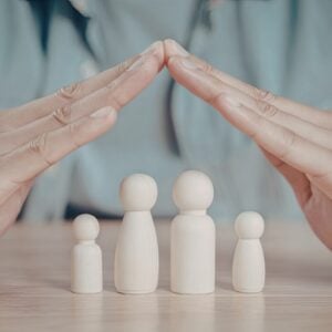 Hands hovering over wooden family figures, forming a protective dome shape over them on a table.