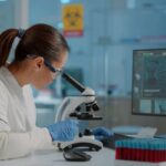 Female scientist in safety goggles and gloves uses a microscope at a lab bench, with a computer monitor and test-tube rack nearby.