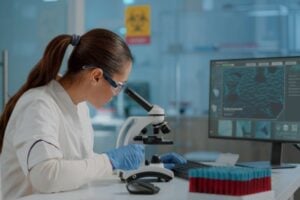 Female scientist in safety goggles and gloves uses a microscope at a lab bench, with a computer monitor and test-tube rack nearby.