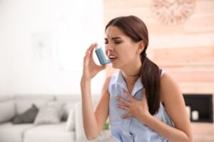 Woman using a blue inhaler to her mouth, pressing it as she struggles to breathe and holds her chest in distress at home.