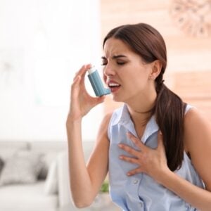 Woman using a blue inhaler to her mouth, pressing it as she struggles to breathe and holds her chest in distress at home.