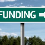 Green directional road sign reading 'FUNDING' with a right-pointing arrow against a blue sky backdrop, suggesting funding to the right.