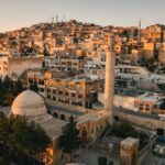 Aerial view of a historic Middle Eastern city at sunset, featuring a domed mosque and a tall minaret among sandstone buildings.